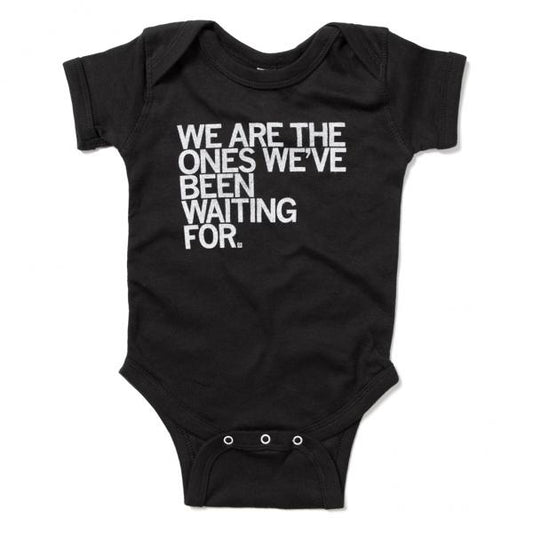Black sweatshop-free infant bodysuit featuring the white printed text, WE ARE THE ONES WEVE BEEN WAITING FOR, inspired by the words of June Jordan.