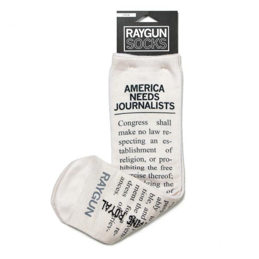 Off-white, American-made crew socks featuring the text AMERICA NEEDS JOURNALISTS and a portion of the First Amendment, with RAYGUN printed on the foot.