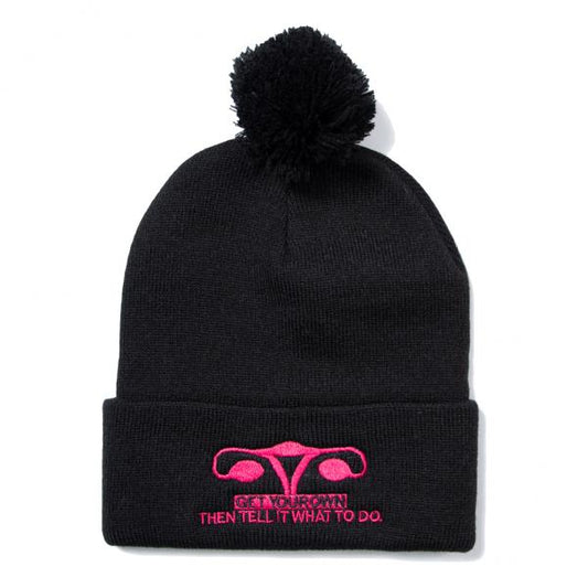 Black pom-pom beanie featuring a neon fuchsia embroidered uterus graphic and the text GET YOUR OWN THEN TELL IT WHAT TO DO.