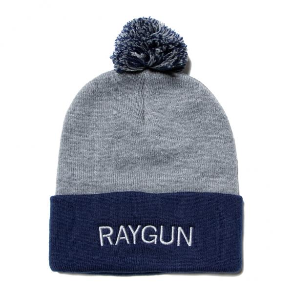 Grey knit beanie with a navy blue folded cuff featuring RAYGUN embroidered in white text and a navy and grey pom-pom on top.