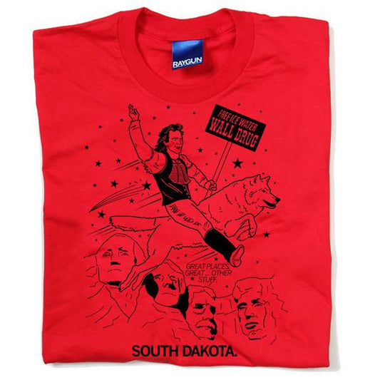 A red Raygun t-shirt with a South Dakota mashup graphic featuring a person riding a wolf, the Wall Drug sign, Mount Rushmore presidential portraits, and the text SOUTH DAKOTA.