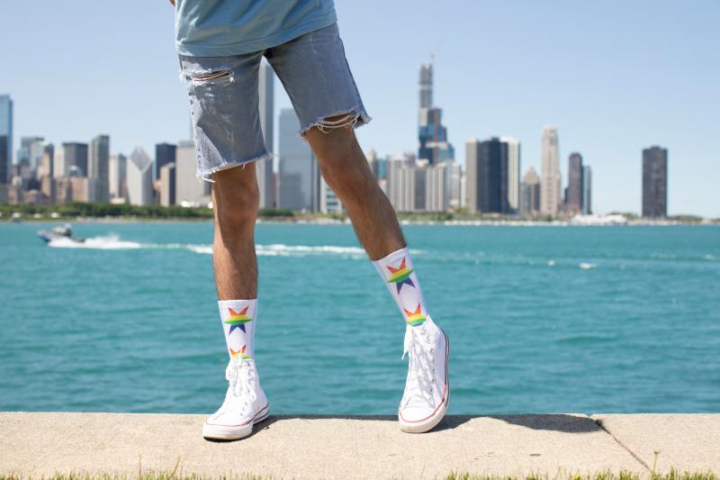 A person wearing denim shorts, white high-top sneakers, and handmade Gay Pride socks with a rainbow star pattern, standing on a pier in front of a city skyline and water.