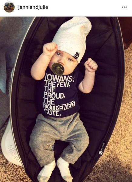 A baby wearing a beanie and a navy blue RAYGUN onesie that reads, Iowans: The Few, The Proud, The Extremely Cute, lying in a black infant seat.