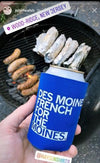 A hand holds a blue scuba foam can cooler with white text reading Des Moines in front of a grill with sausages and oysters.