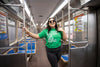 A smiling woman wearing a green Heather Kelly t-shirt that says I train Spanish for the train stands in the center of an empty subway car, holding onto a metal pole.