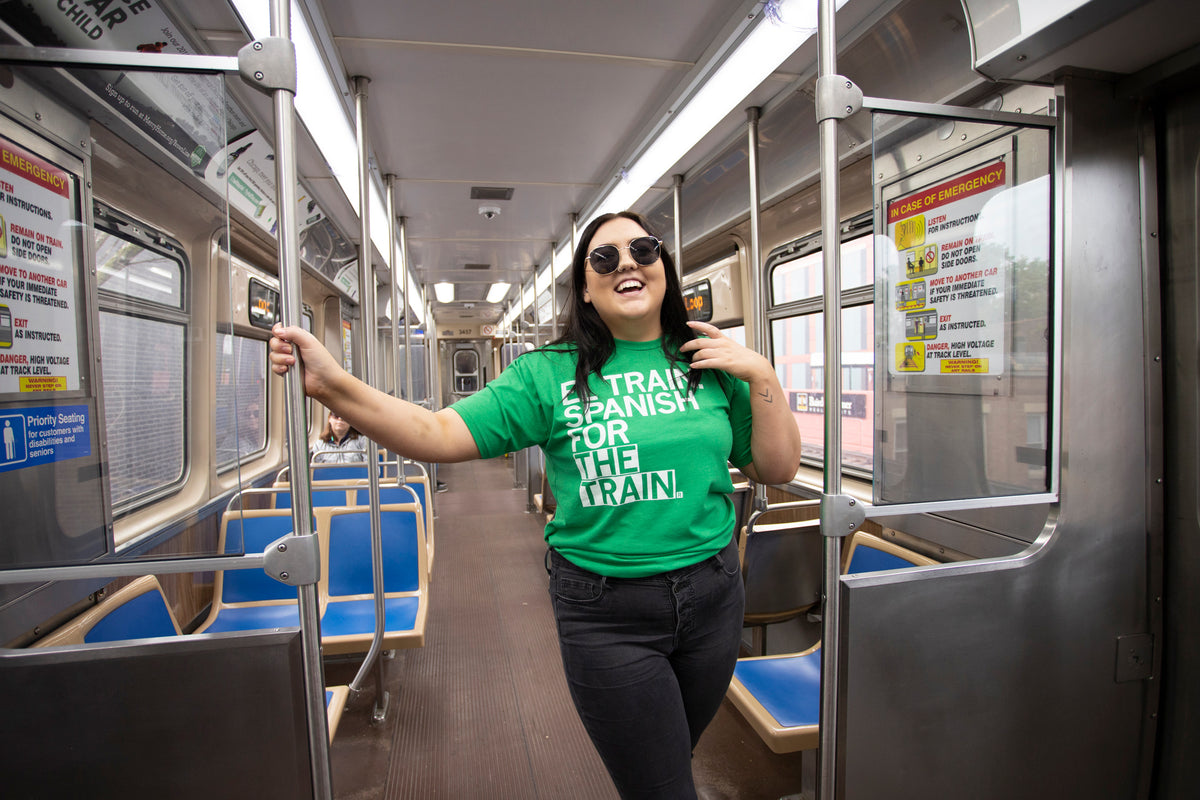 A smiling woman wearing a green Heather Kelly t-shirt that says I train Spanish for the train stands in the center of an empty subway car, holding onto a metal pole.