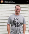 A smiling man wearing glasses and a heather grey Busch Light t-shirt stands in front of a white wall.