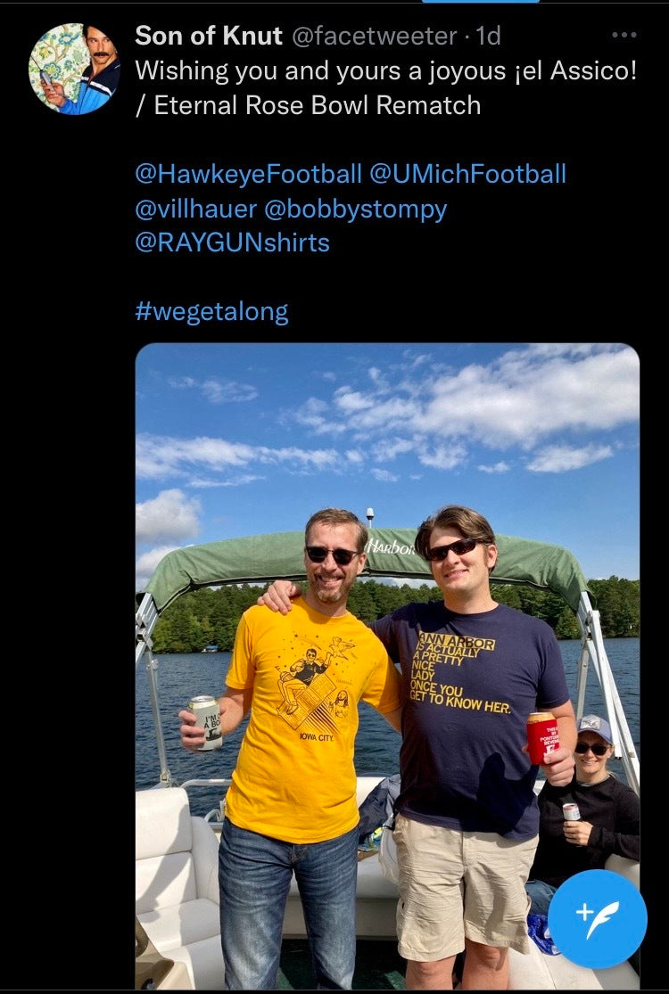 Two men stand on a boat on a lake; one wears an Iowa City yellow t-shirt, and the other wears a blue t-shirt that says Ann Arbor is actually a pretty nice lady once you get to know her, both shirts being union made.