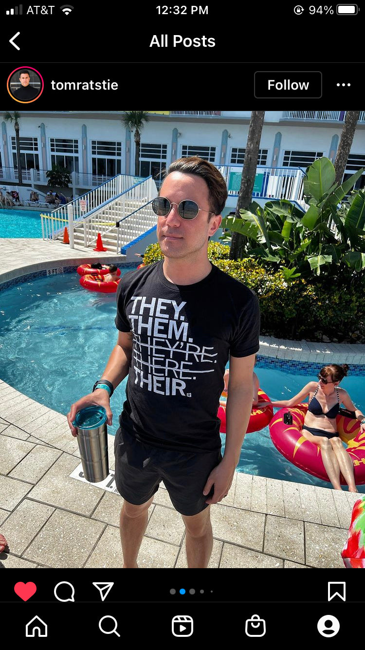 A man wearing sunglasses and a gender pronoun t-shirt that reads THEY. THEM. THEYRE. HERE. THEIR. stands by a swimming pool.