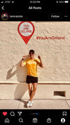 A man wearing a union made, gold HAPPY t-shirt and sunglasses stands in front of a wall with a YOU ARE DELAND sign and the hashtag #YouAreDeland.
