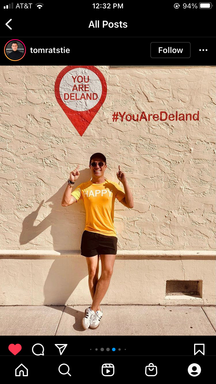A man wearing a union made, gold HAPPY t-shirt and sunglasses stands in front of a wall with a YOU ARE DELAND sign and the hashtag #YouAreDeland.