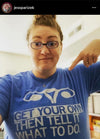 A woman points to her blue, union-made t-shirt featuring an illustration of a uterus and the text, GET YOUR OWN THEN TELL IT WHAT TO DO.