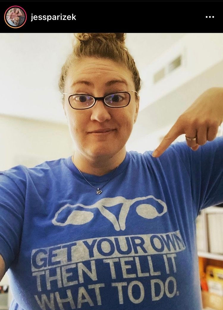 A woman points to her blue, union-made t-shirt featuring an illustration of a uterus and the text, GET YOUR OWN THEN TELL IT WHAT TO DO.