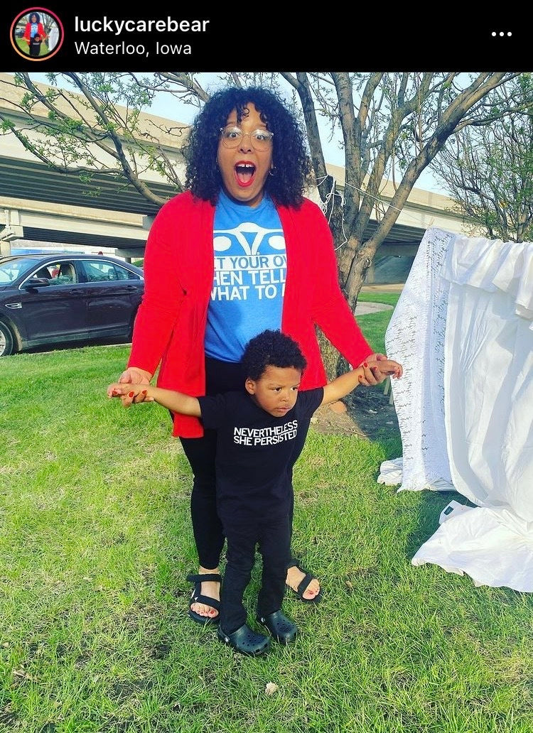 A woman in a red cardigan and blue Planned Parenthood t-shirt stands behind a young child in a black t-shirt that reads Nevertheless She Persisted, both wearing union labor apparel with their arms outstretched.