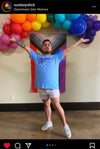 A person wearing a light blue RAYGUN t-shirt that says Probably Still Gay, standing in front of a Progress Pride flag and an arch of rainbow balloons.