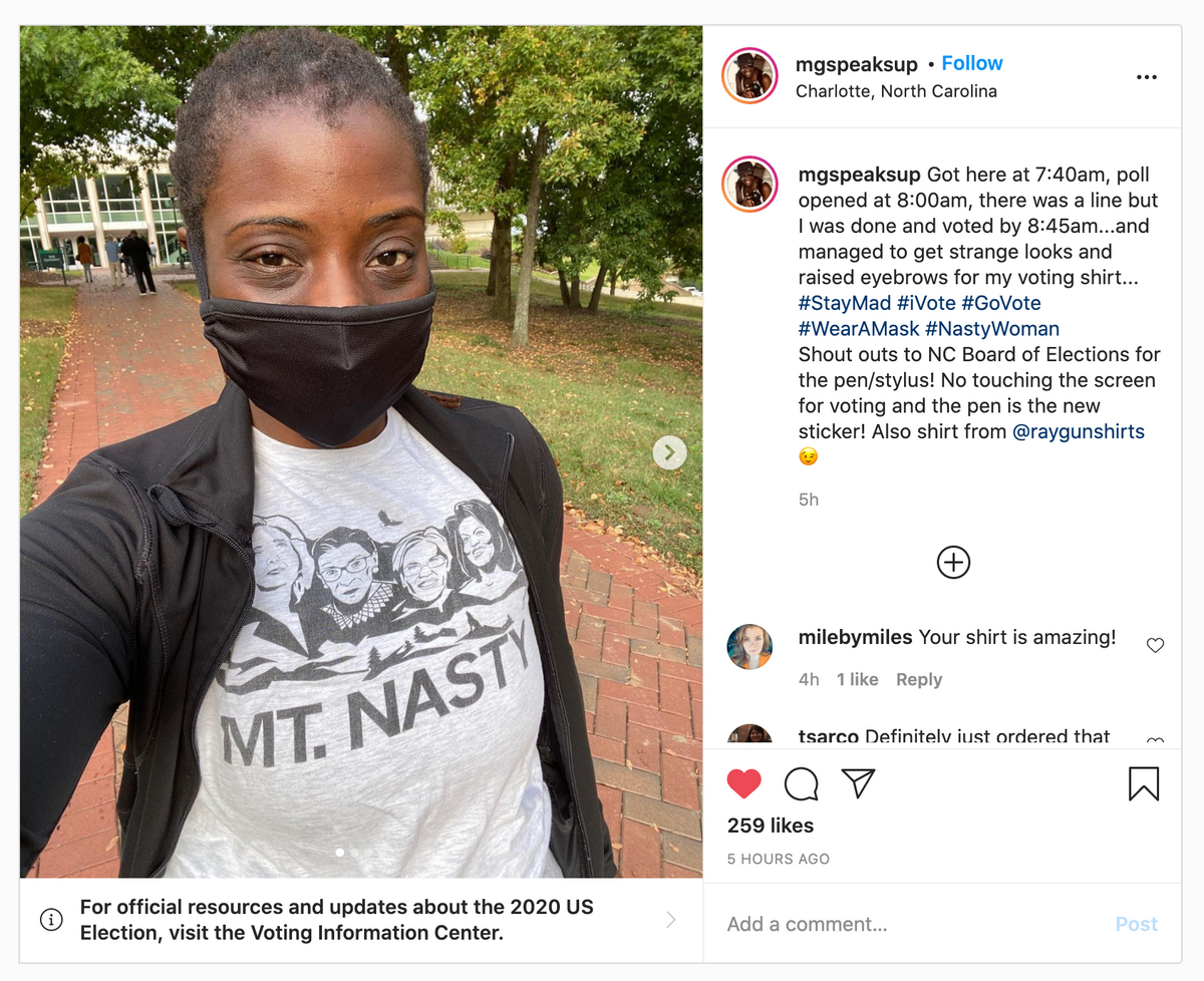 A person wearing a black mask and a Nasty Women t-shirt featuring the faces of four women and the text MT. NASTY stands on a brick path outside a polling place.