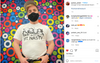 A man wearing a black face mask and a Raygun Nasty Women t-shirt stands in front of a colorful, circular patterned wall.