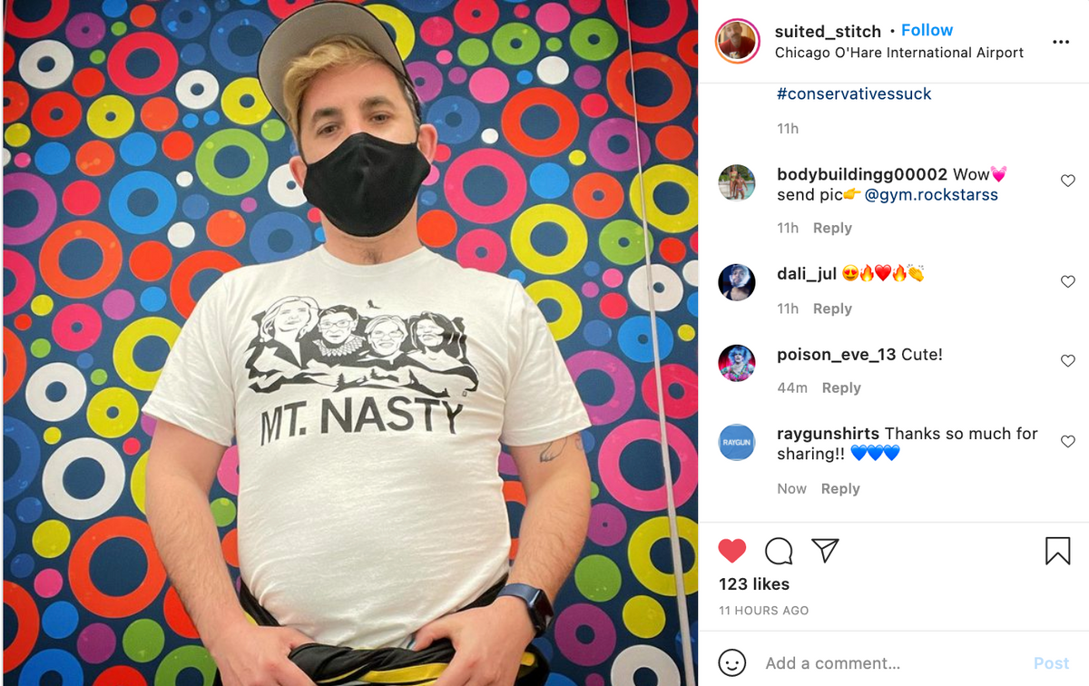 A man wearing a black face mask and a Raygun Nasty Women t-shirt stands in front of a colorful, circular patterned wall.