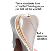 A hand holding an open RAYGUN notebook to show its lay-flat binding and labelable spine, with text explaining: These notebooks have a lay-flat binding so you can fold all the way flat and a spine you can label.