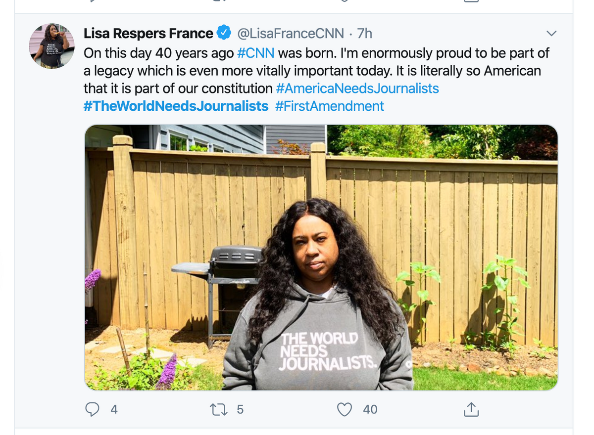 A woman stands in front of a wooden fence wearing a union made pullover hoodie that reads, The world needs journalists.