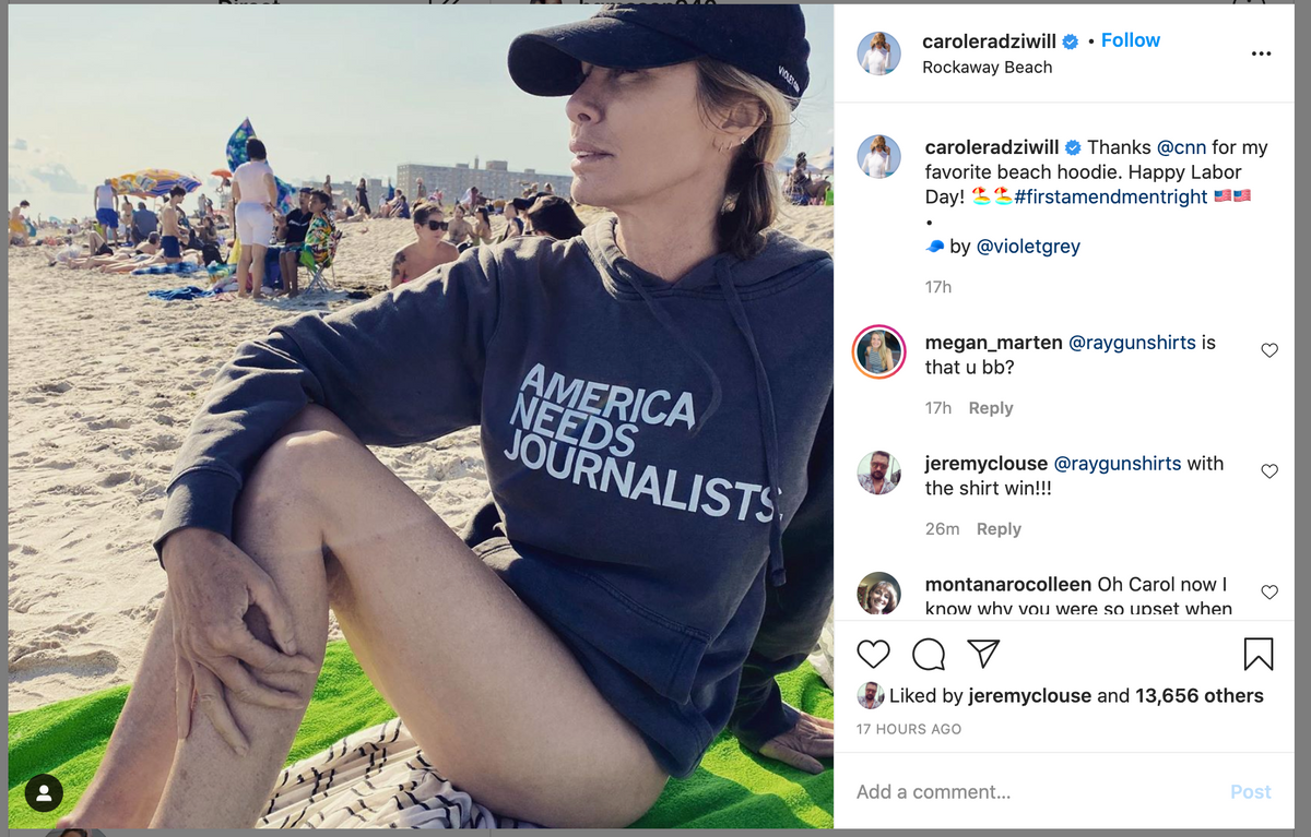 A woman wearing a black baseball cap and a Pigment Black Pullover Hoodie that reads America Needs Journalists sits on a beach towel on a crowded beach.