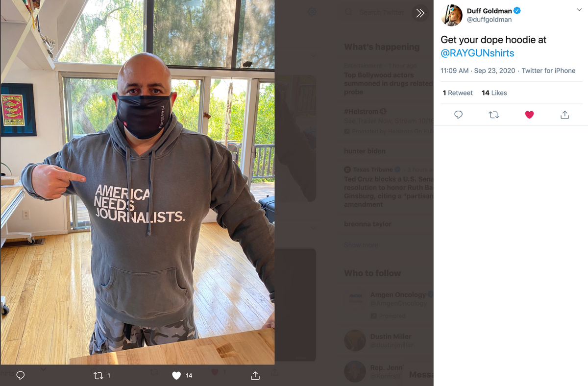 A person wearing a face mask and a Pigment Black Pullover Hoodie that says America Needs Journalists points to the text on the garment.