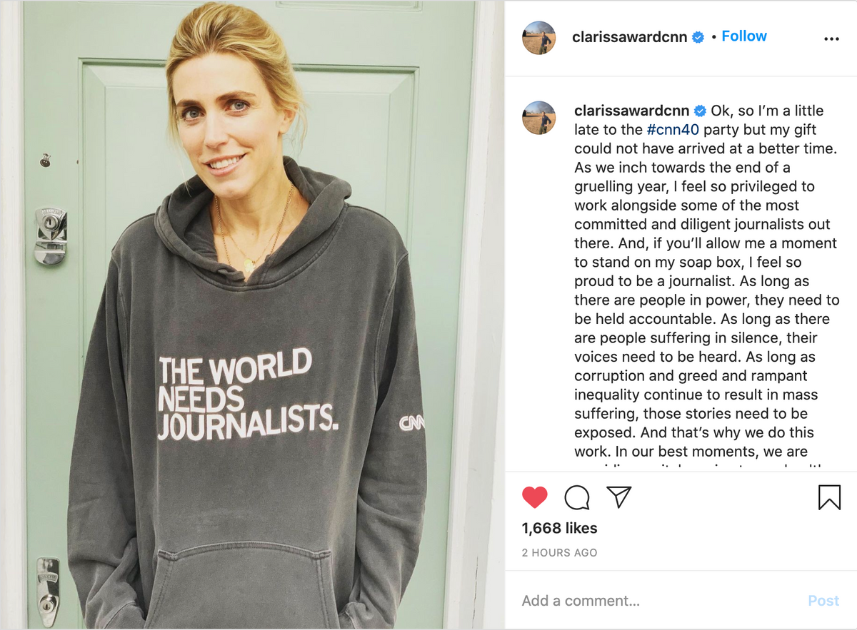 A woman wearing a union made, dark grey pullover hoodie that reads THE WORLD NEEDS JOURNALISTS with a small CNN logo on the sleeve.