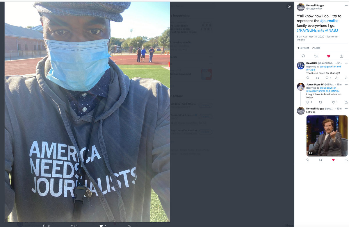 A man wearing a blue face mask, a flat cap, and an America Needs Journalists pigment black pullover hoodie stands on a track field.