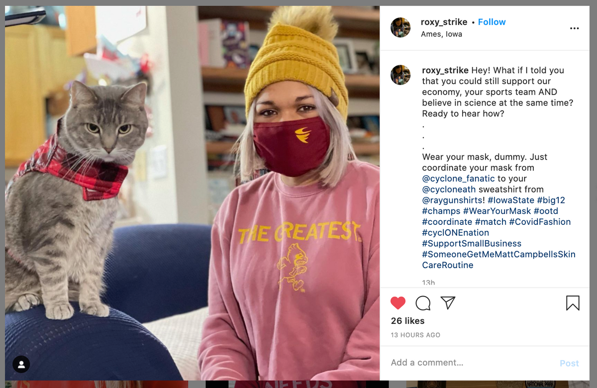 A woman in a yellow beanie and a maroon RAYGUN sweatshirt face mask with a gold screen print sits next to a grey tabby cat wearing a matching maroon bandana.