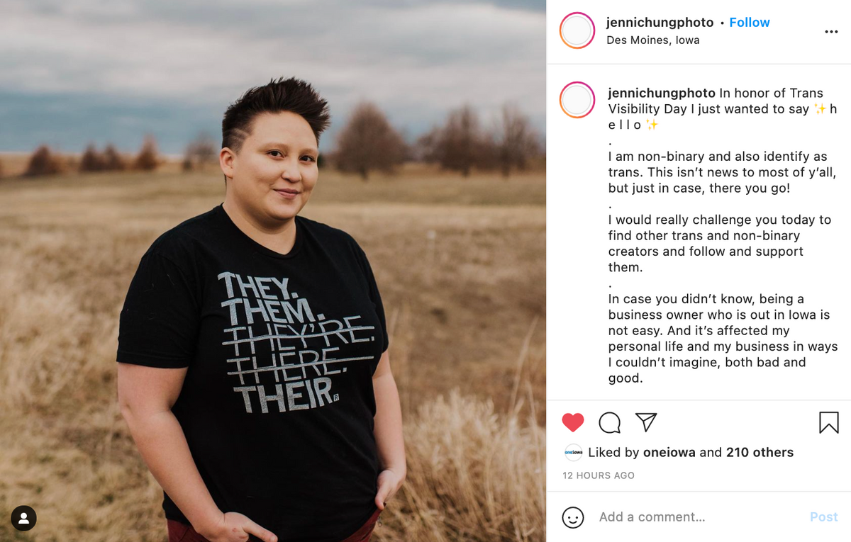 A person with a short haircut stands in a field wearing a sweatshop-free black t-shirt printed with the words THEY. THEM. THEYRE. THERE. THEIR.