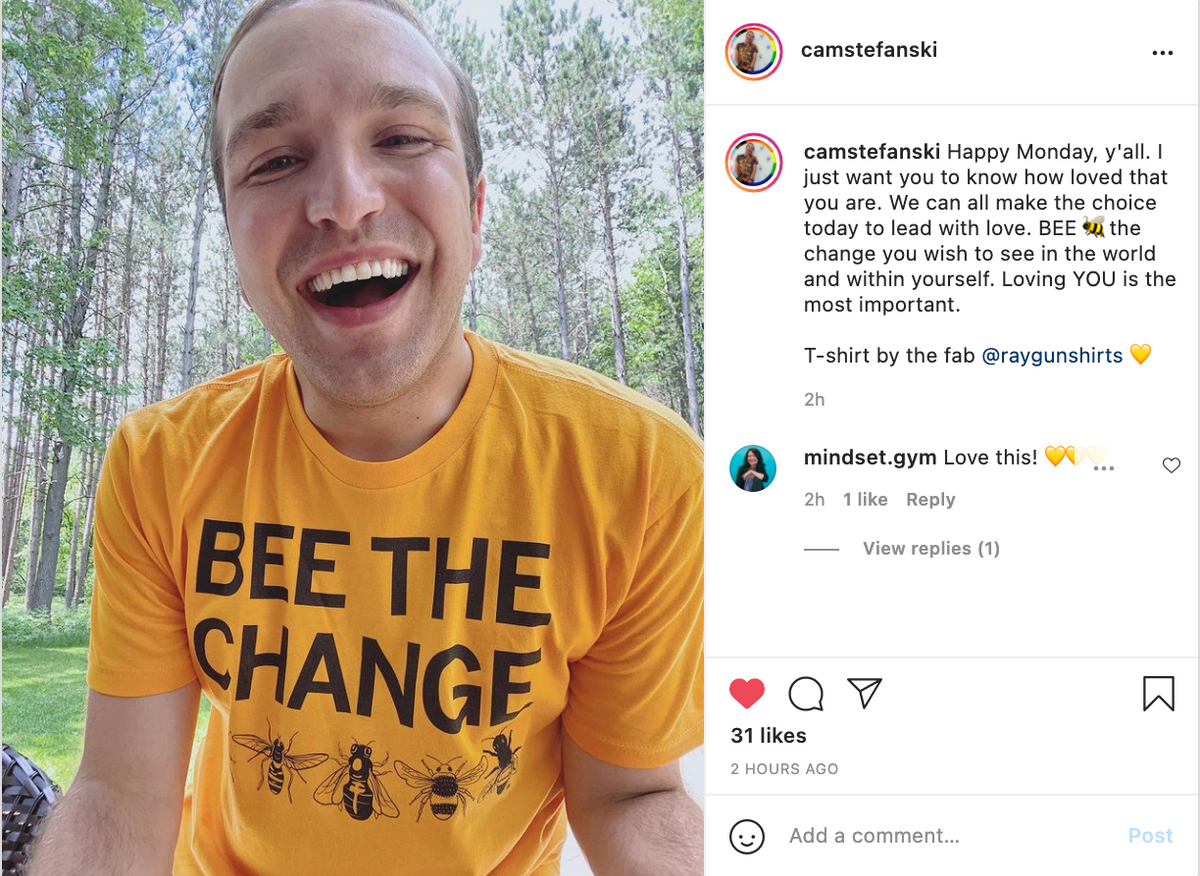 A person smiling at the camera wearing a yellow, union-made RAYGUN T-shirt that says BEE THE CHANGE with illustrations of bees.