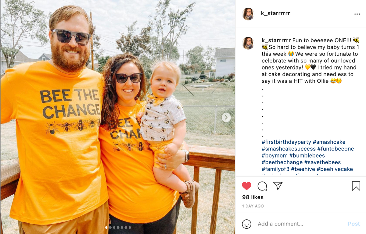A family of three wears matching RAYGUN bee-themed shirts while standing on a wooden deck, with a father holding a baby in a yellow outfit next to a smiling mother.