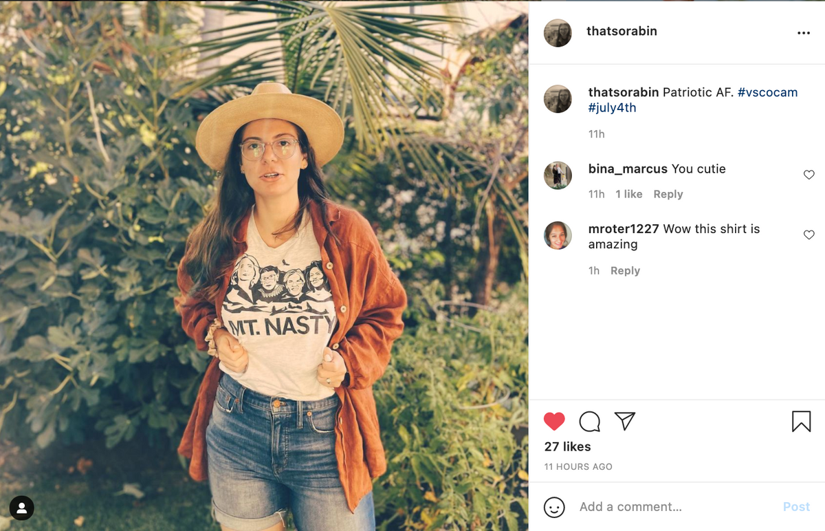A person wearing a wide-brimmed hat, glasses, and a Raygun t-shirt that reads MT. NASTY with a graphic of faces, standing outdoors in front of plants.