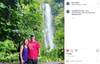 A man wearing an America Needs Public Schools t-shirt and a woman in a purple tank top stand together in front of a waterfall surrounded by lush tropical greenery.