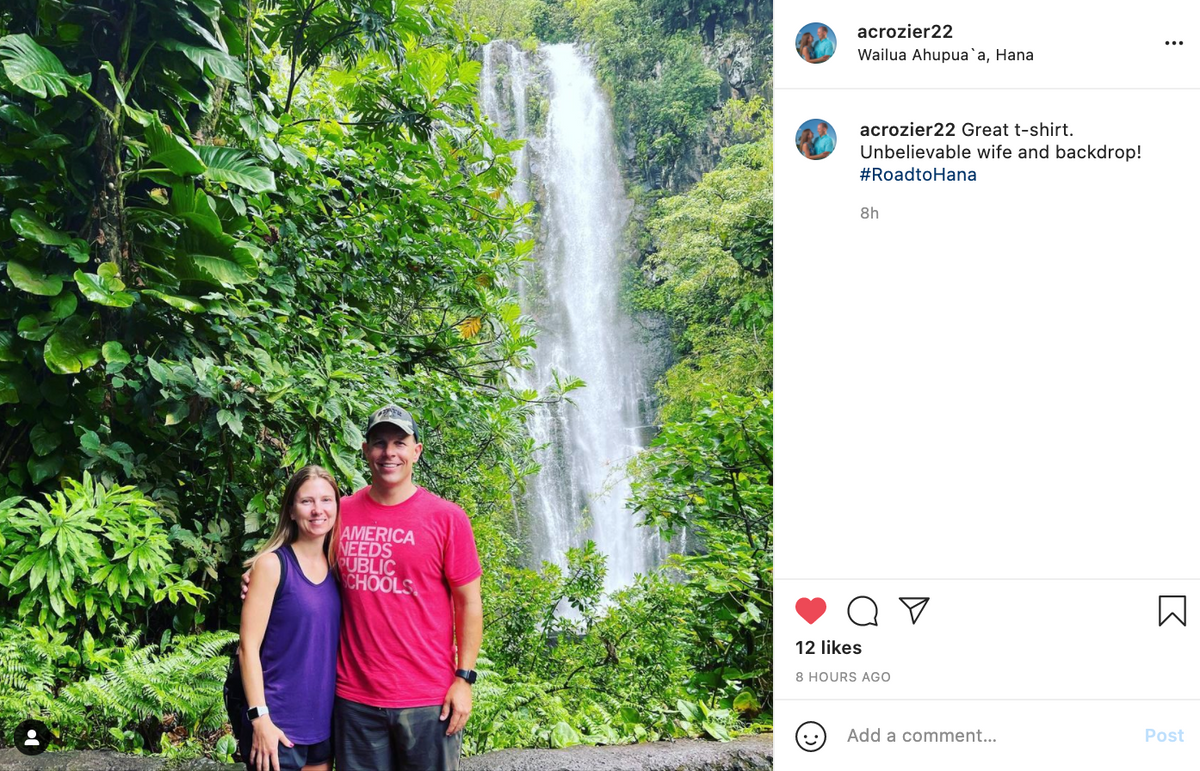 A man wearing an America Needs Public Schools t-shirt and a woman in a purple tank top stand together in front of a waterfall surrounded by lush tropical greenery.