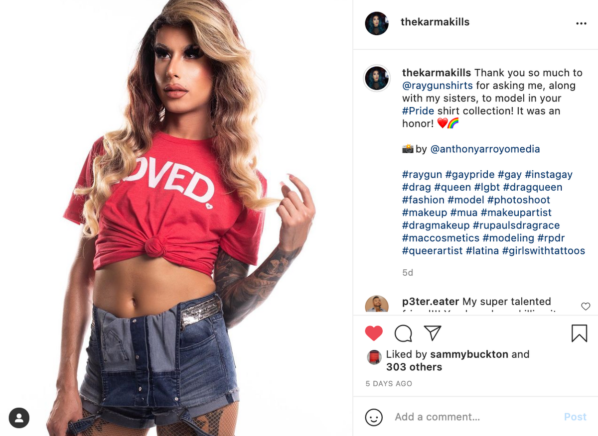 A drag queen with long wavy blonde hair poses wearing a red union-made t-shirt that says LOVED and denim shorts.