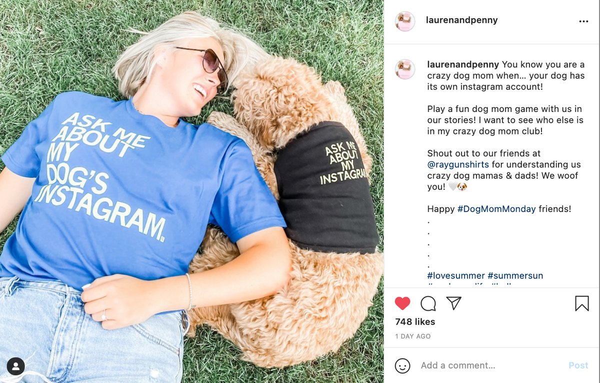 A woman wearing a blue RAYGUN dog shirt that says Ask me about my dogs Instagram lies in the grass next to a golden dog wearing a black vest that also says Ask me about my Instagram.