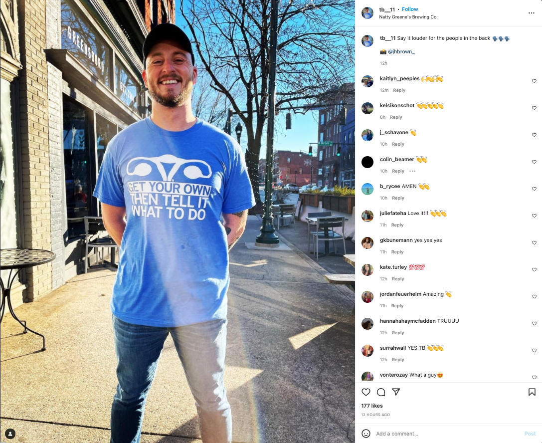 A man wearing a union labor blue t-shirt that says Get your own then tell it what to do with a graphic of a uterus stands on a sidewalk in front of a Planned Parenthood building.