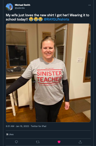 A woman wears a heather grey t-shirt that says Just another sinister teacher over a black long-sleeved shirt.