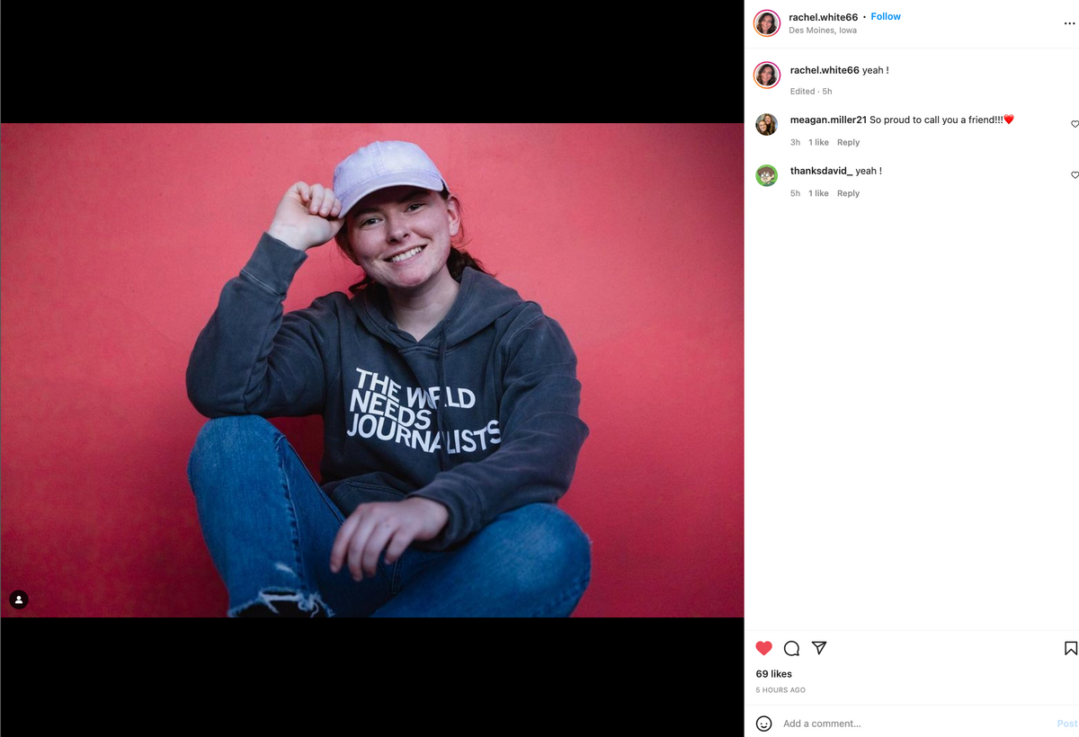 A smiling person wears a light purple baseball cap and a grey America Needs Journalists hoodie while sitting against a red wall.