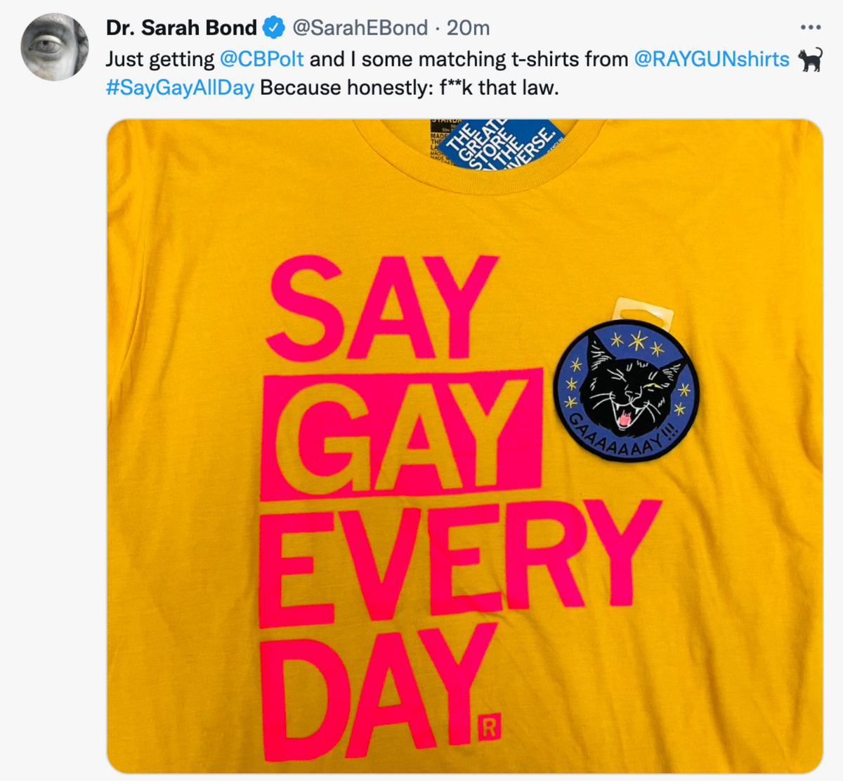 A yellow supportive pride apparel t-shirt with SAY GAY EVERY DAY in large pink font, featuring a circular black pin of a cat saying GAAAAAAAY!!! and a tag from THE GREATEST STORE IN THE UNIVERSE.