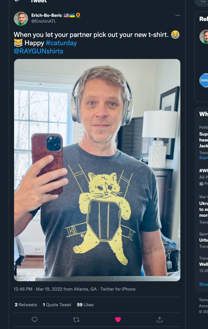 A man wearing a charcoal black RAYGUN t-shirt with a graphic of a cat sitting on a stool takes a mirror selfie.