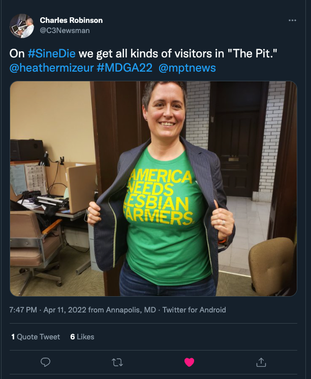 A person smiles while wearing an open blazer over a heather kelly green t-shirt that reads AMERICA NEEDS LESBIAN FARMERS.
