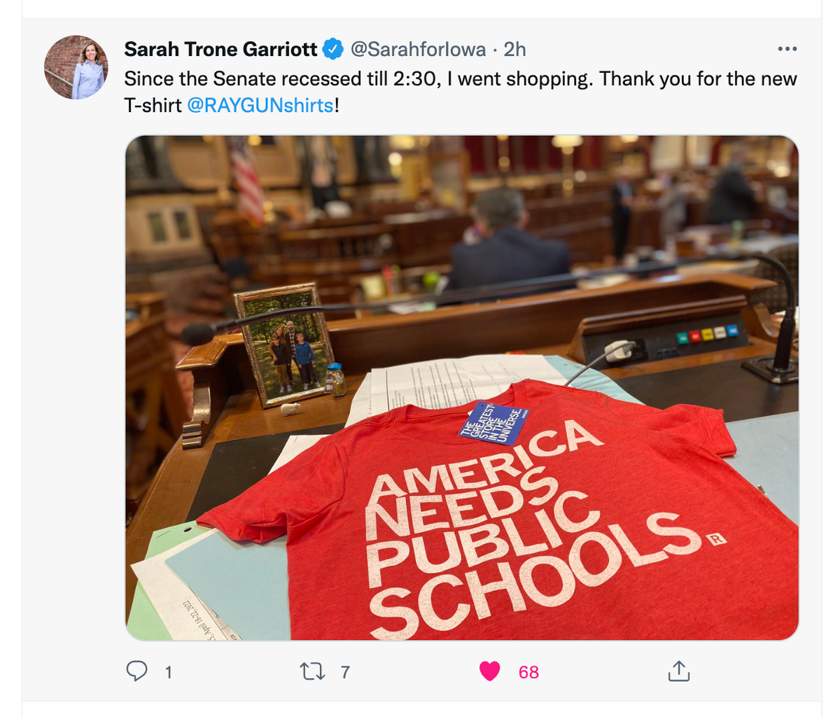 A red union-made t-shirt printed with AMERICA NEEDS PUBLIC SCHOOLS rests on a wooden desk inside a legislative chamber.