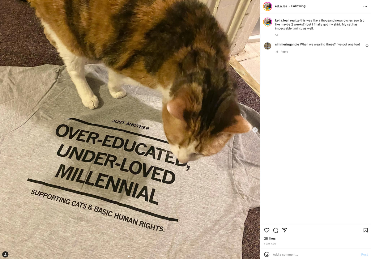 A calico cat stands on a gray, union made millennial t-shirt printed with the text, Just another over-educated, under-loved millennial, supporting cats & basic human rights.