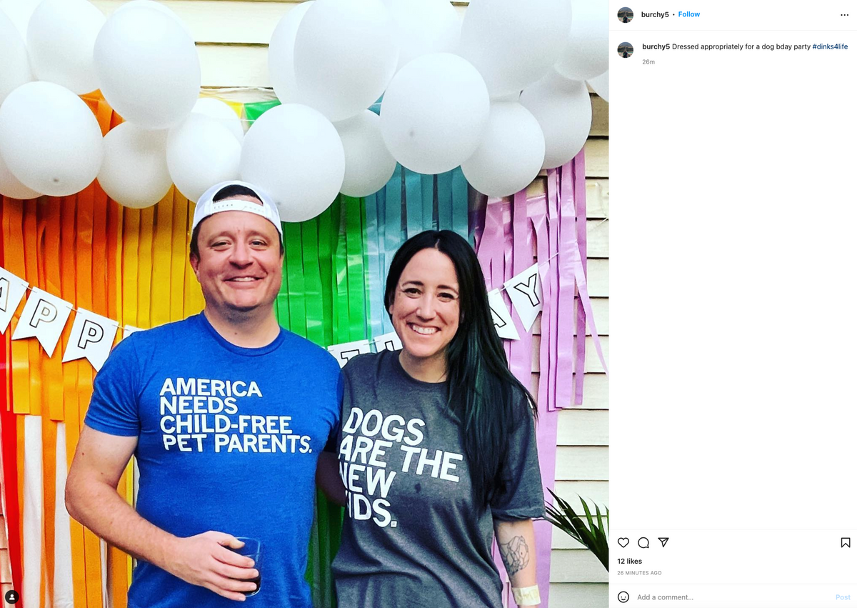 A man in a union made blue t-shirt and a woman in a grey t-shirt stand in front of a rainbow streamer backdrop decorated with white balloons and a Happy Birthday banner.
