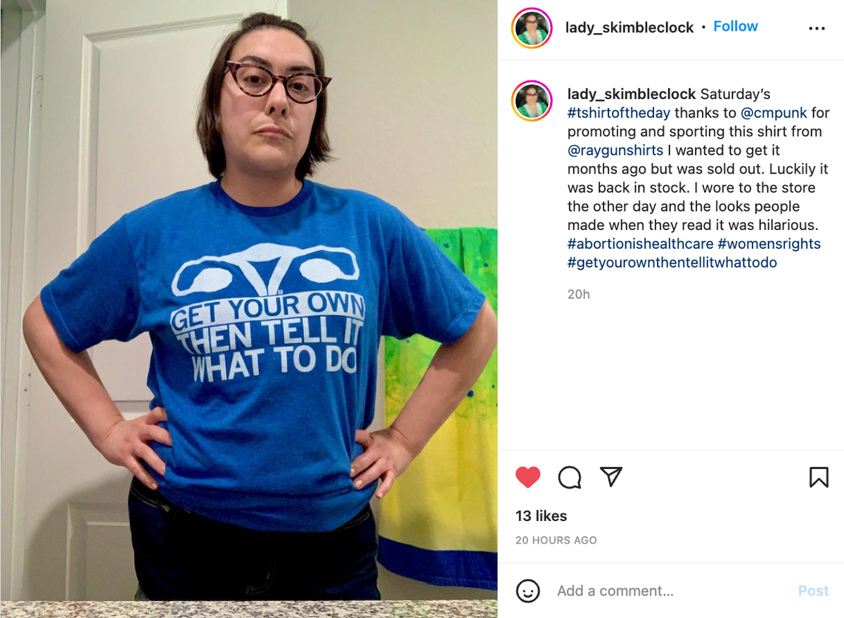 A person with short dark hair and glasses wears a sweatshop-free blue t-shirt featuring a stylized uterus graphic and the text GET YOUR OWN THEN TELL IT WHAT TO DO.