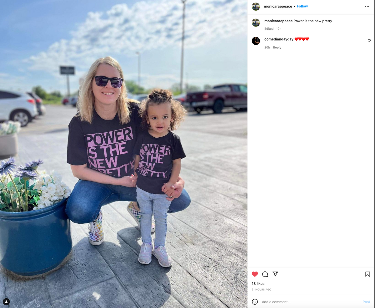 A woman and a young child crouch outdoors wearing matching black T-shirts that say Power Is The New Pretty in pink letters.