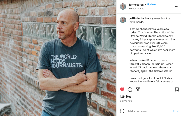 A man with crossed arms leans against a brick wall, wearing a union-made t-shirt that reads, THE WORLD NEEDS JOURNALISTS.