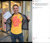 A smiling man holds a mail-in ballot and opens his jacket to reveal an I Voted sticker and a yellow LGBTQ+ t-shirt that says Say gay every day.